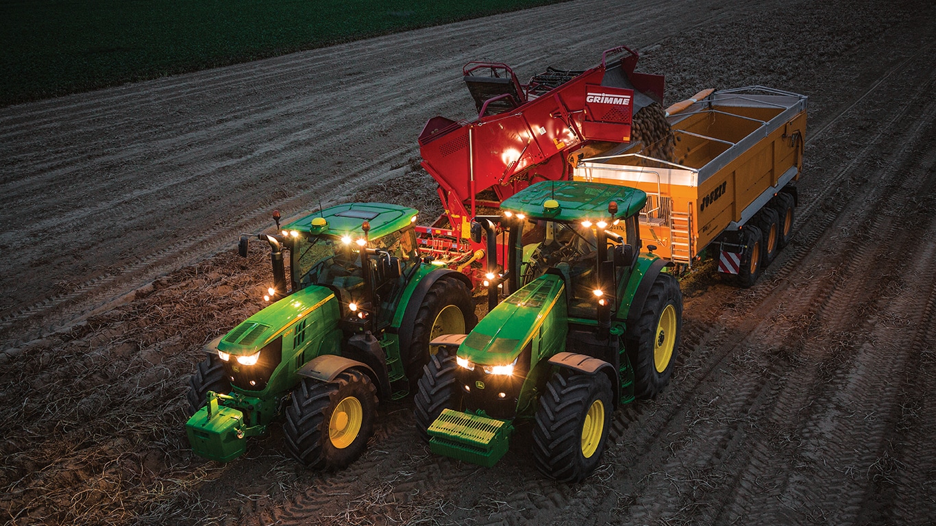 Traktor, der nachts Kartoffelernte-Maschine zieht, entl&auml;dt Kartoffeln in einen Anh&auml;nger, der von einem anderen Traktor gezogen wird