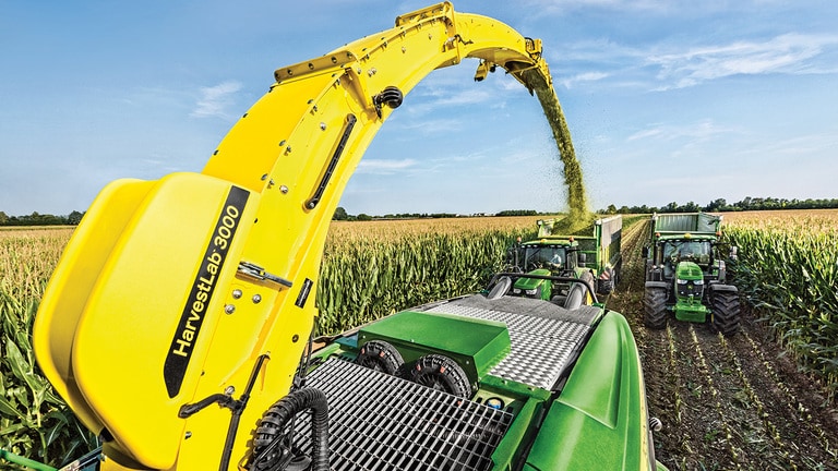 Blick von hinten auf den selbstfahrenden Feldh&auml;cksler der Serie 9000 von John Deere mit zwei Silage-Wagen, die von John Deere-Traktoren direkt dahinter gezogen werden