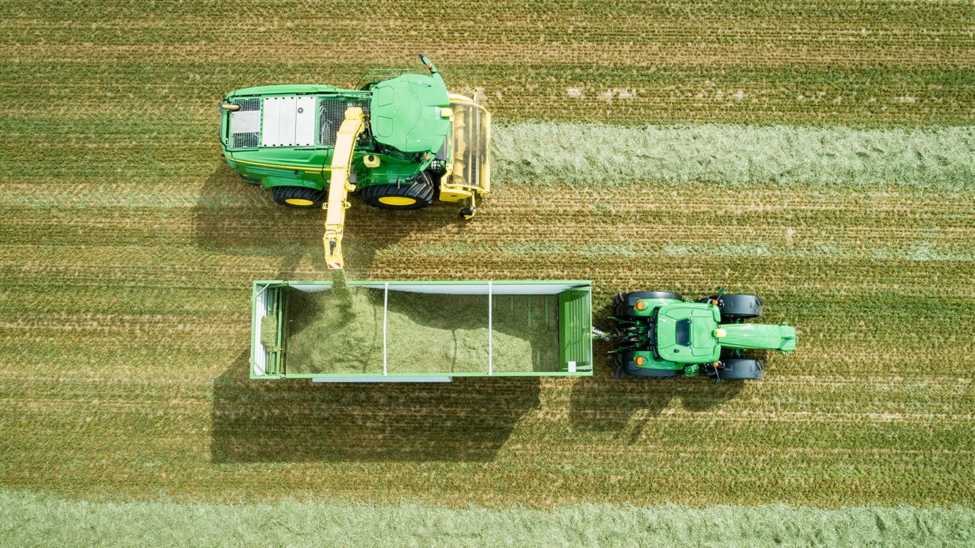 Luftbild eines selbstfahrenden Feldh&auml;cksler der John Deere 8000-Serie mit Gras-Pickup, der Silage in einen Wagen l&auml;dt, der von einem John Deere-Traktor gezogen wird