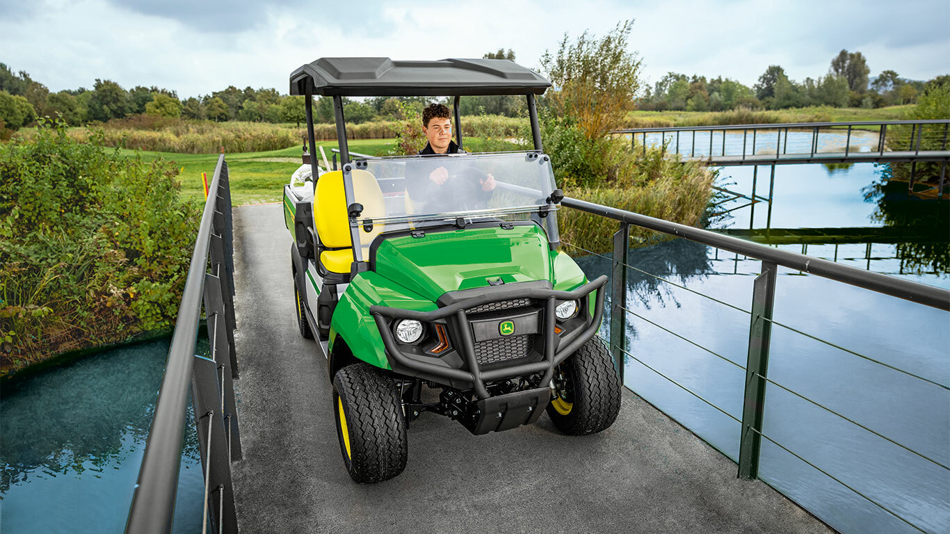 John Deere Gator GS Nutzfahrzeug überquert eine schmale Brücke über ein ruhiges Gewässer, umgeben von üppigem Grün und Bäumen. John Deere Gator GS Nutzfahrzeug überquert eine schmale Brücke über ein ruhiges Gewässer, umgeben von üppigem Grün und Bäumen.