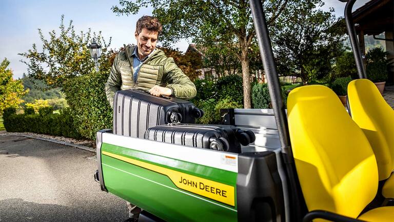 Eine Person lädt einen großen schwarzen Koffer in die Ladefläche eines John Deere Gator GS mit leuchtend gelben Sitzen, umgeben von Grün und Bäumen.