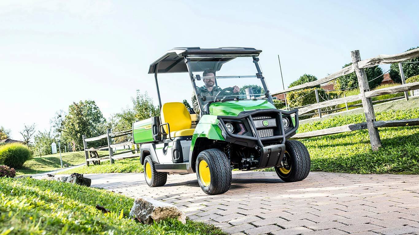 Grünes John Deere Gator GS Nutzfahrzeug fährt auf einem befestigten Weg durch eine gestaltete Grünanlage mit Rasen und Holzzäunen.