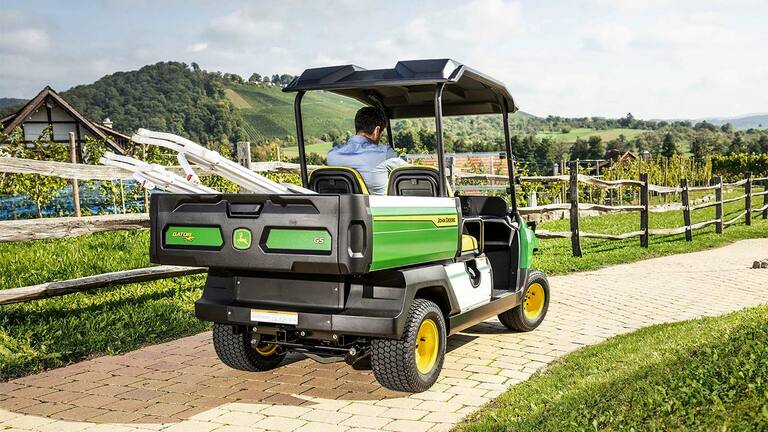Rückansicht eines John Deere Gator GS, der einen gepflasterten Weg entlangfährt, mit malerischen Hügeln und Weinbergen im Hintergrund.