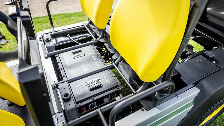 Nahaufnahme von zwei Lithium-Batteriepacks, die unter den leuchtend gelben Sitzen eines John Deere Gator GS Electric Nutzfahrzeugs installiert sind.