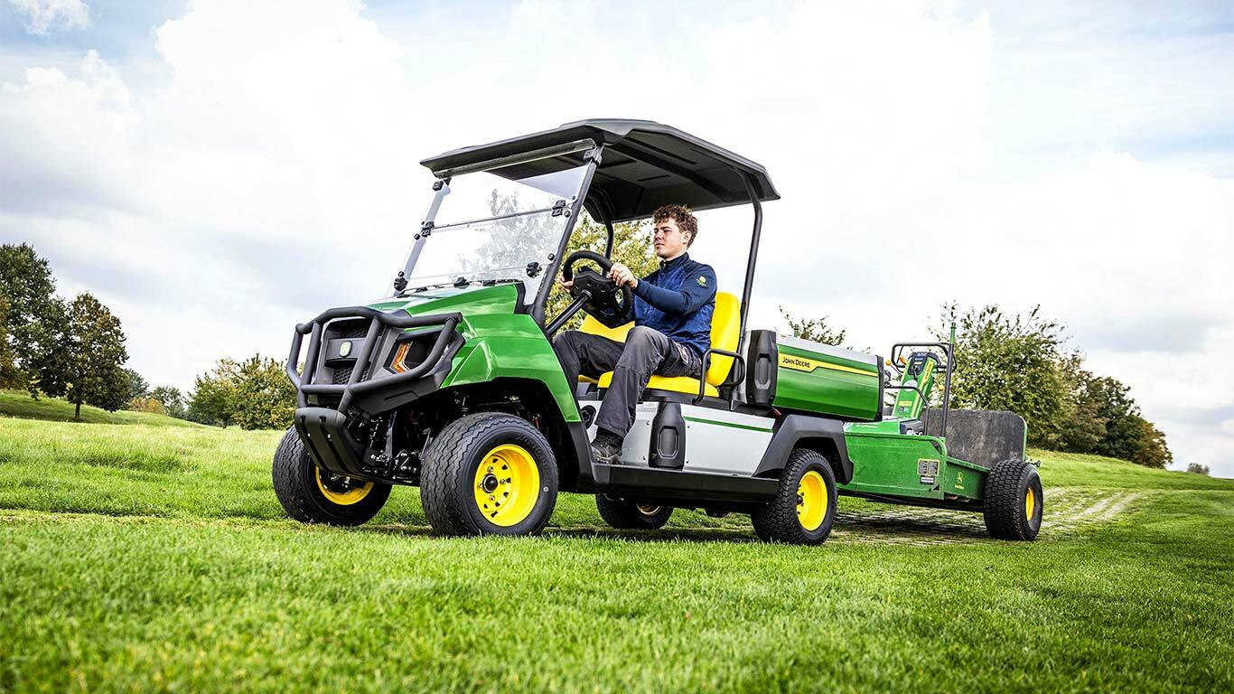 Grünes John Deere Gator Electric Nutzfahrzeug zieht Geräte über ein grasbewachsenes Feld unter teilweise bewölktem Himmel.