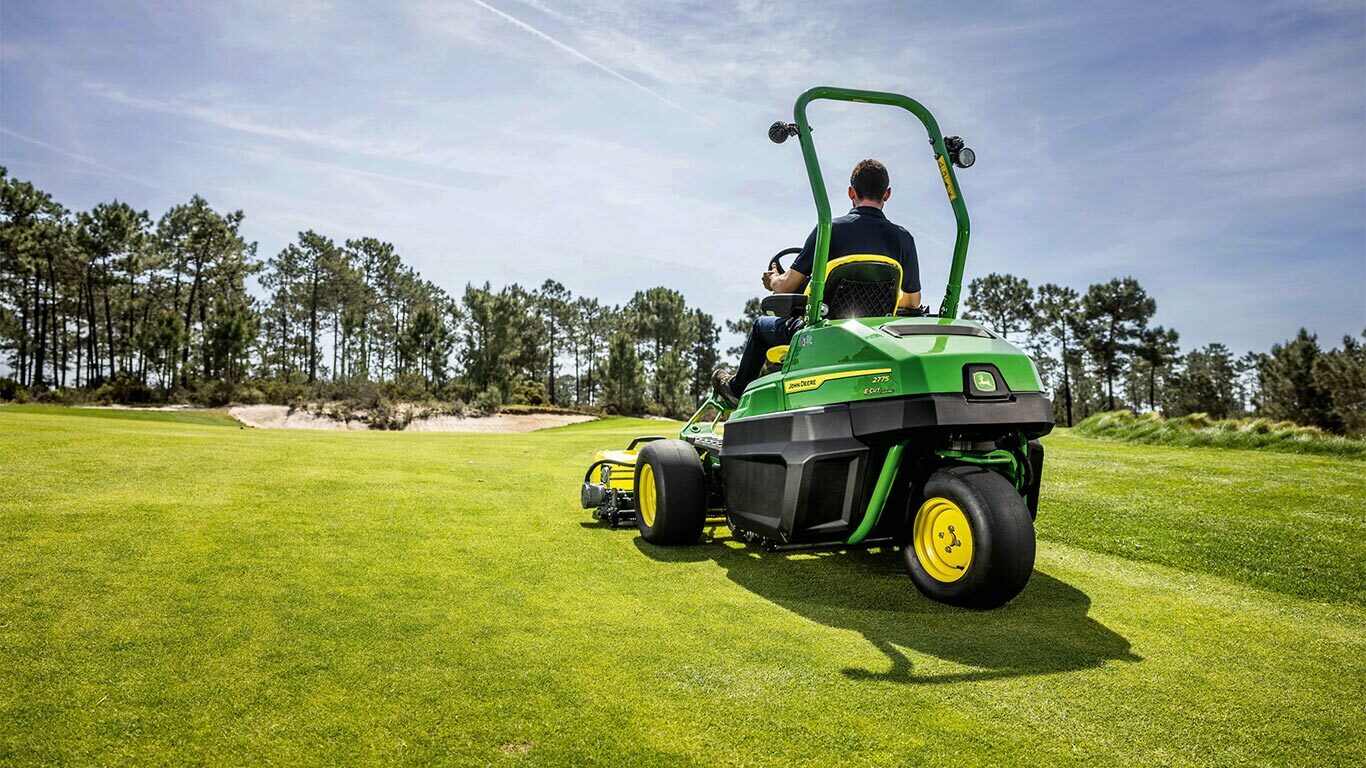 Eine Person fährt mit einem grünen John Deere 2775 E-Cut™ Greens-Elektromäher über einen gepflegten Golfplatz mit Bäumen und einem teilweise bewölkten Himmel im Hintergrund.