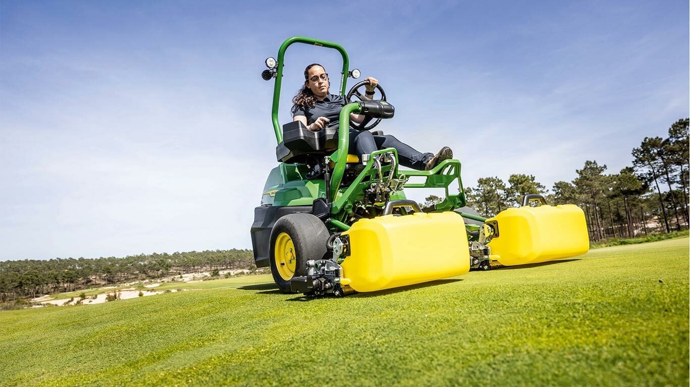 Eine Person bedient auf einem Golfgrün mit Bäumen und einem klaren blauen Himmel im Hintergrund einen grün-gelben John Deere 2775 E-Cut™ Elektromäher mit Vorlaufrollen, die von einem gelben Gehäuse abgedeckt sind.