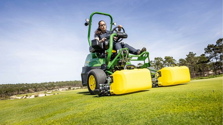 Eine Person bedient auf einem Golfgrün mit Bäumen und einem klaren blauen Himmel im Hintergrund einen grün-gelben John Deere 2775 E-Cut™ Elektromäher mit Vorlaufrollen, die von einem gelben Gehäuse abgedeckt sind.