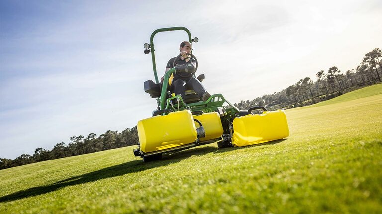 Eine Person bedient auf einem Golfgrün mit Bäumen und einem teilweise bewölkten Himmel im Hintergrund einen grün-gelben John Deere 2775 E-Cut™ Elektromäher mit drei großen Vorlaufrollen, die von gelben Gehäusen abgedeckt sind.