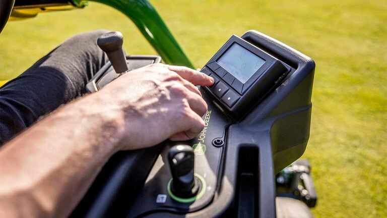 Nahaufnahme der Hand einer Person, die das TechControl-Bedienfeld an einem John Deere 2775 E-Cut™ Elektromäher steuert und Schaltflächen auf einer Digitalanzeige mit einem Joystick und anderen sichtbaren Bedienelementen drückt.