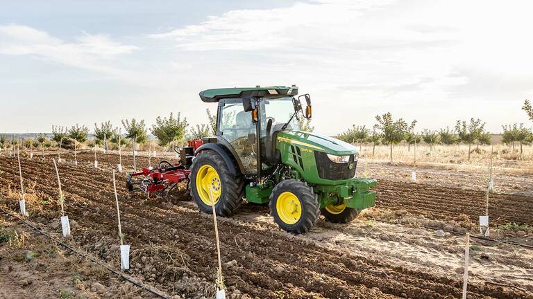 John Deere 5075EN Traktor bearbeitet den Boden auf einem offenen Feld mit klarem Himmel und gepflanzten Reihen.