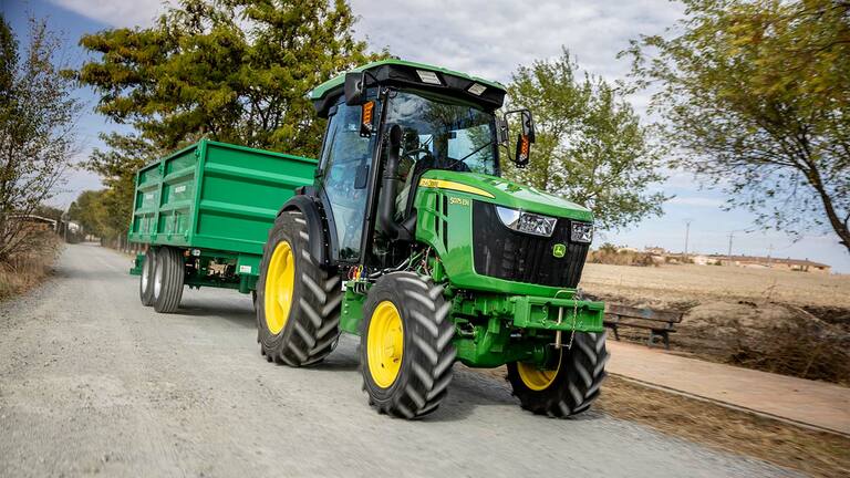 John Deere 5075EN Traktor zieht einen grünen Anhänger auf einer ländlichen Schotterstraße mit Bäumen im Hintergrund.
