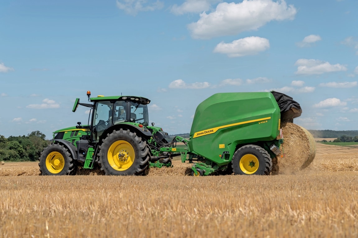 John Deere V452R auf einem Strohfeld, gezogen von einem John Deere 6R 150, beim Auswerfen eines Ballens