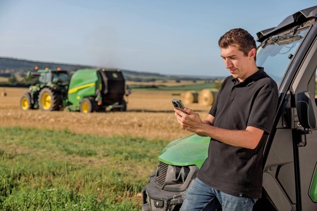 Person, die die Daten der Ballendokumentation auf dem Mobiltelefon überprüft, mit einer John Deere Ballenpresse VR im Hintergrund