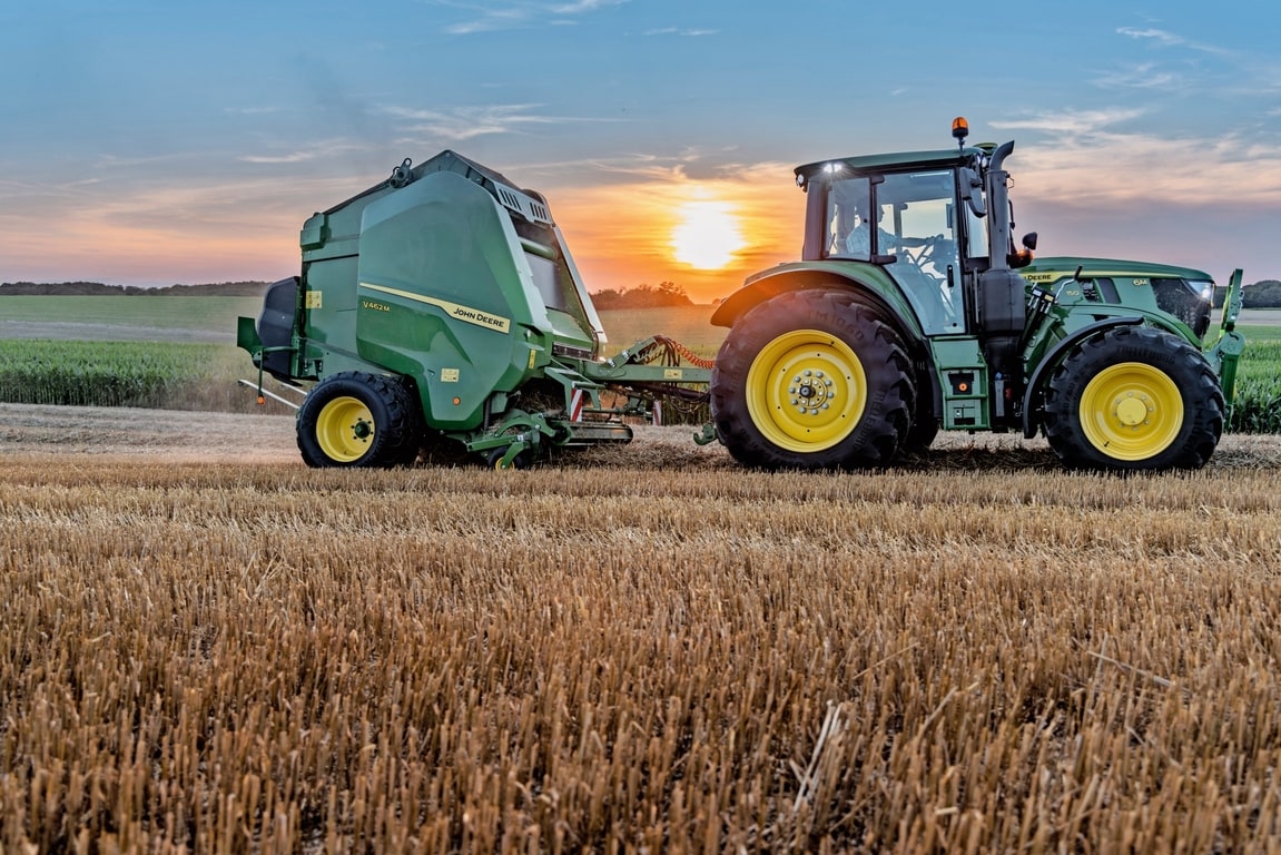 John Deere V462M auf einem Strohfeld, gezogen von einem John Deere 6M 150 bei Sonnenuntergang