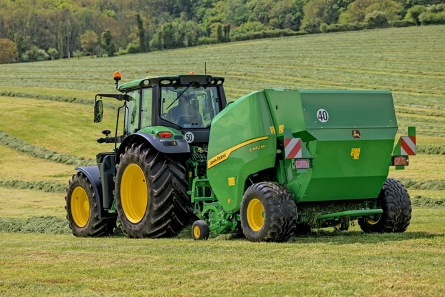 John Deere F442M Ballenpresse, gezogen von einem John Deere 6M Traktor auf einer hügeligen Wiese