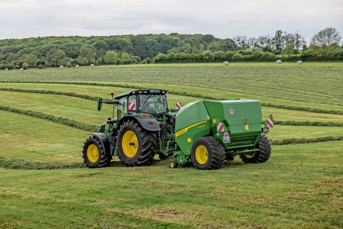 John Deere Ballenpresse F442R wird von einem John Deere 6M Traktor auf einem hügeligen Grasfeld gezogen